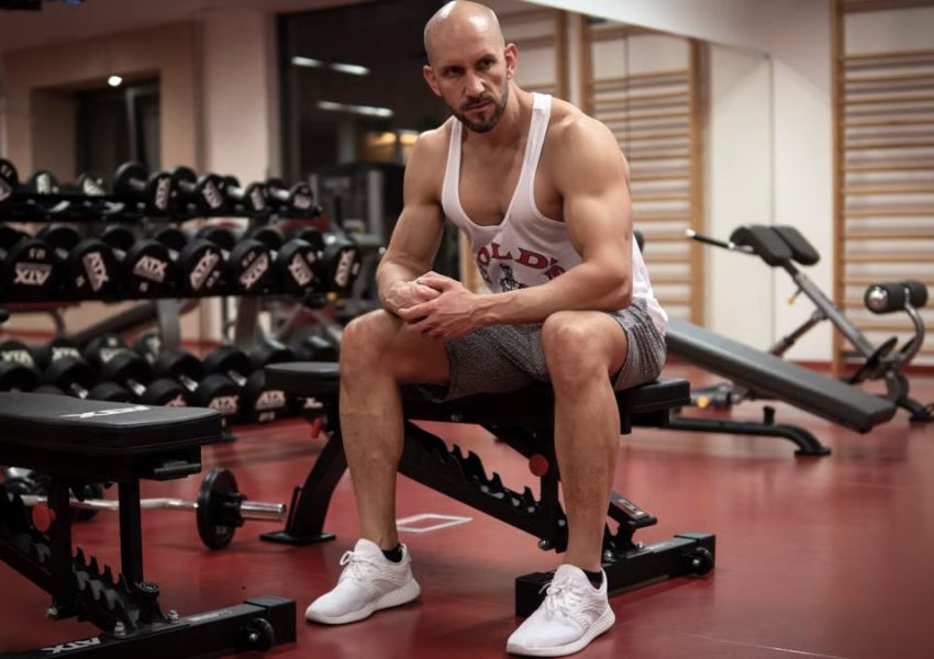 Strong, determined man in white tank top sitting in gym, showcasing fitness and strength.